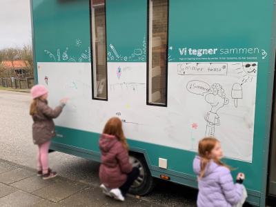 Bogbilen holder ved et fortov. Tre børn er i gang med at tegne på den.