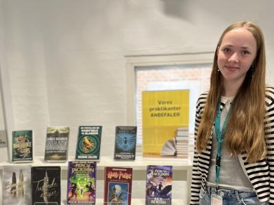 Viola står ved siden af sin udstilling med gode bøger, som hun anbefaler. Viola kigger direkte ind i kameraet og smiler.