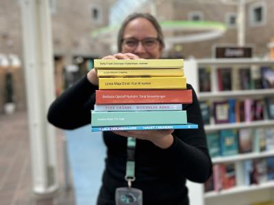 Tine står blandt fyldte bogreoler på Hovedbiblioteket. Hun holder syv af de omtalte bøger i denne artikel frem for sig i en stak med bogryggene frem. Der er fokus på bogryggene, mens både Tine og resten af baggrunden, som viser biblioteksrummet, fremstår let sløret. Man kan ane, at Tine smiler.