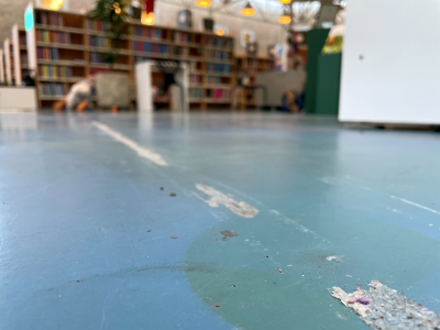 Et halvklat og slidt linoliumsgulv i frøperspektiv. I baggrunden aner man bogreoler og børn på biblioteket.