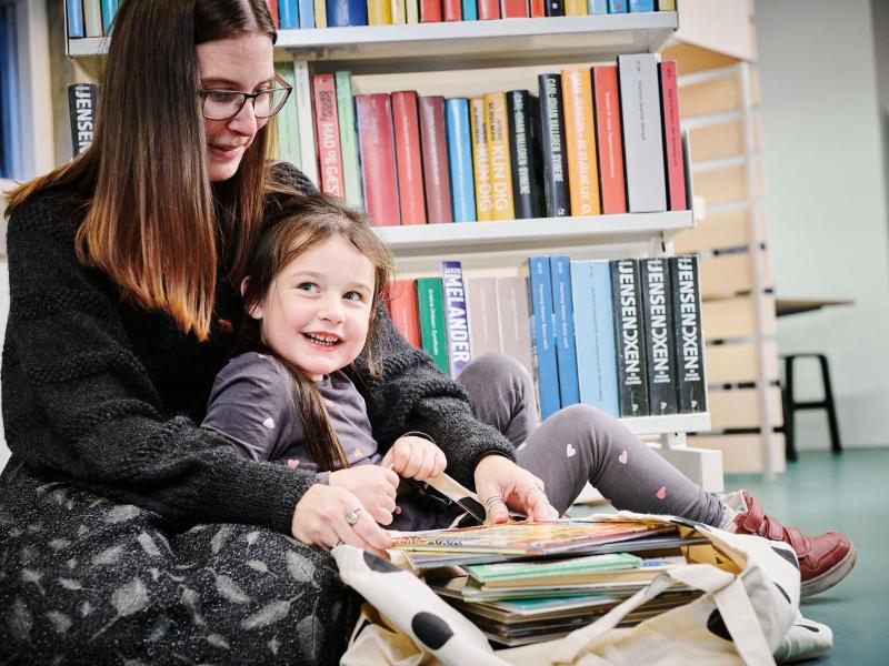 En voksen og et barn sidder i et bibliotek og kigger i en bog sammen