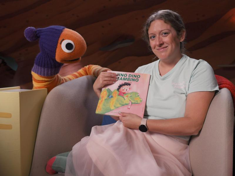 En perso sidder i en blød stol og viser bogen Fino Dino Bambino frem. Til venstre for sidder en stor dukkeudgave af Orm fra DR Minisjangs Børsteuniver. Billedet stammer fra "Læs med Orm"