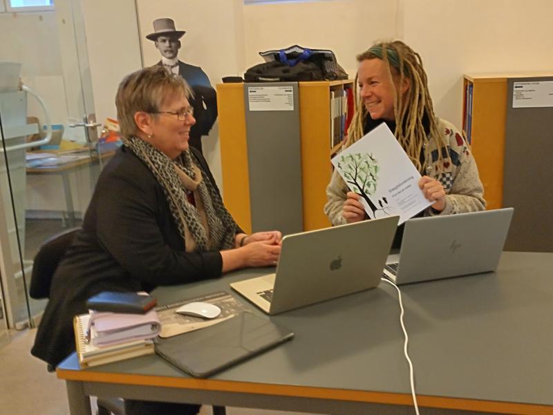 En bibliotekar hjælper en person med slægtsforskning på HistorieAalborg. De sidder begge ved et bord med computere og bøger foran sig. De smiler til hinanden.