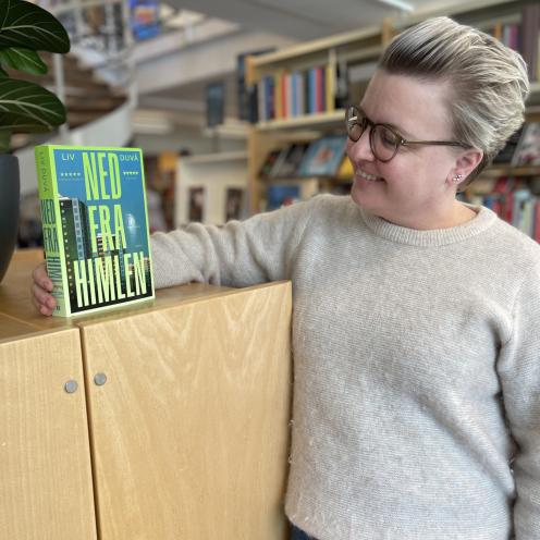Kirstine står på biblioteket. Hun står lænet op af et skab, hvorpå Liv Duvås roman "Ned fra himlen" står med forsiden frem. Kirstine kigger smilende på bogen. Bag hende ses biblioteksrummet med fyldte bogreoler.