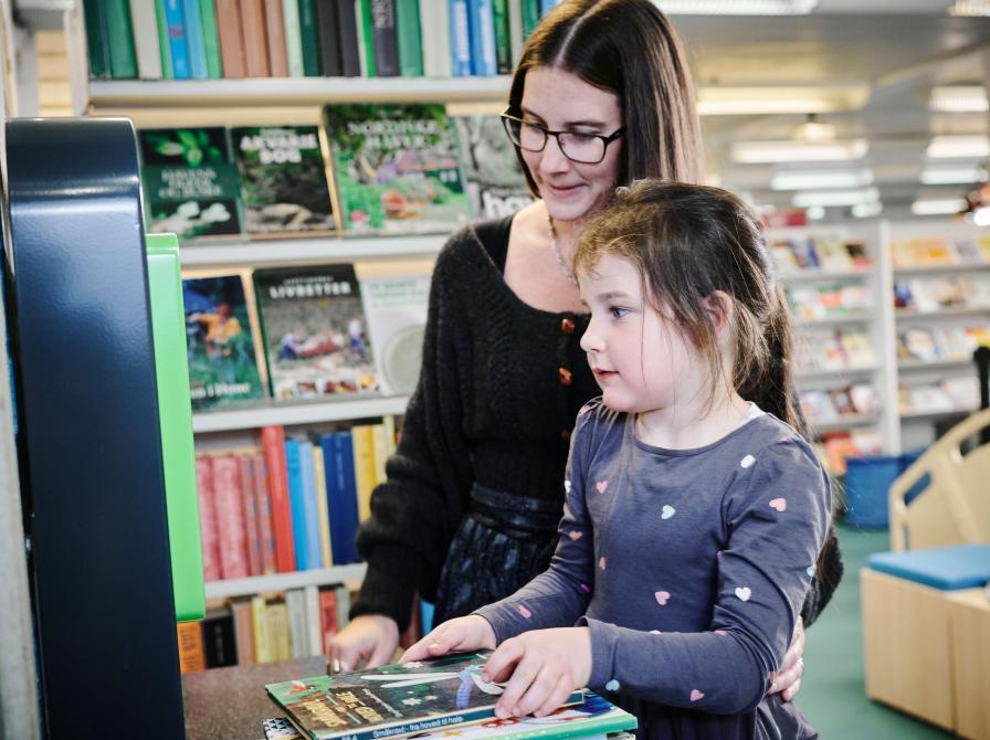 En voksen og et barn står i et bibliotek foran en udlånsautomat