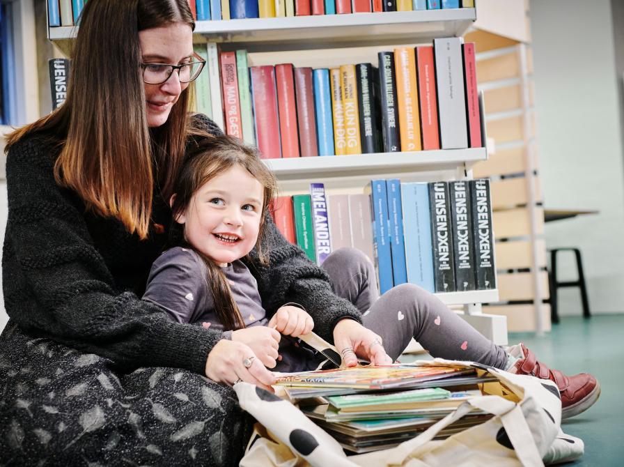 En voksen og et barn sidder i et bibliotek og kigger i en bog sammen