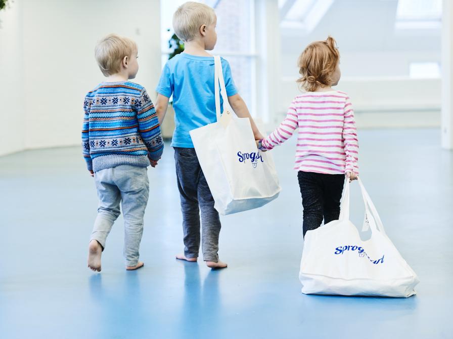 Tre børn går ved siden af hinanden med hinanden i hænderne. To af dem har en sprogstartsæt over skulderen.