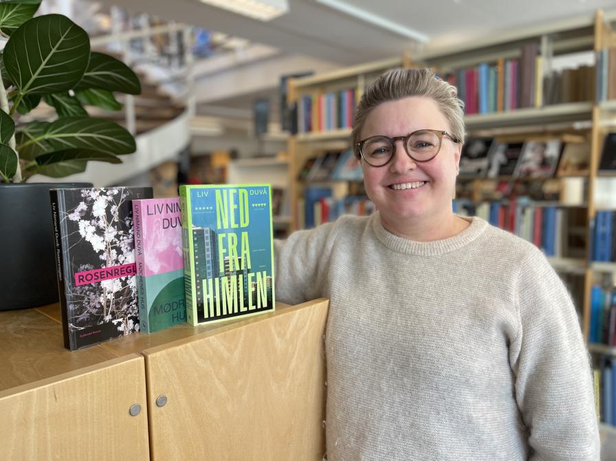 Kirstine står på biblioteket. Hun står lænet op af et skab, hvorpå tre af Liv Duvås romaner står med forsiderne frem. Kirstine smiler og bag hende ses biblioteksrummet med fyldte bogreoler.