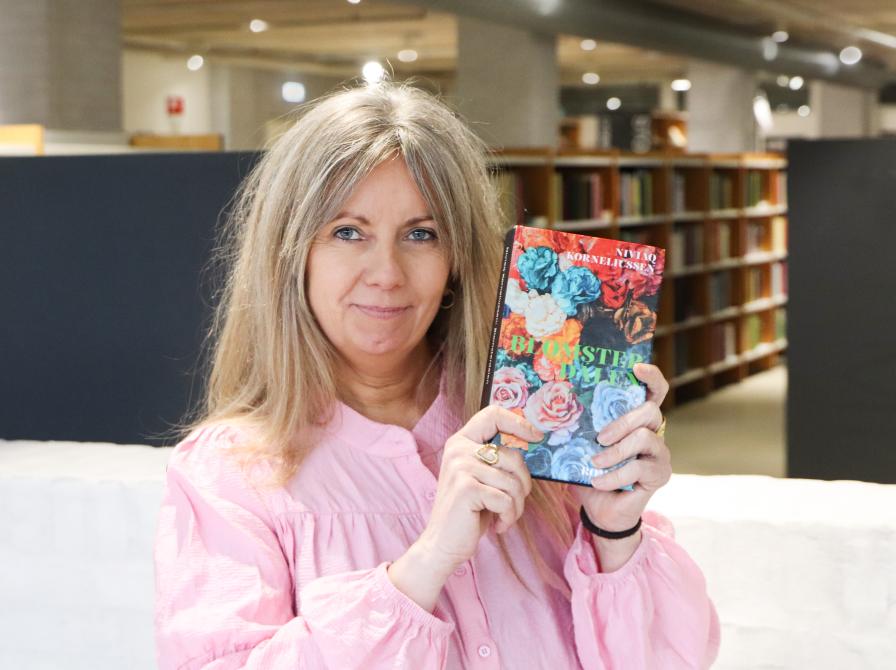 Merete holder bogen "Blomsterdalen" op ved siden af sit hoved, så bogforsiden ses tydeligt. Bag Merete ses biblioteksrummet med fylde bogreoler. Merete kigger direkte ind i kameraet og smiler.