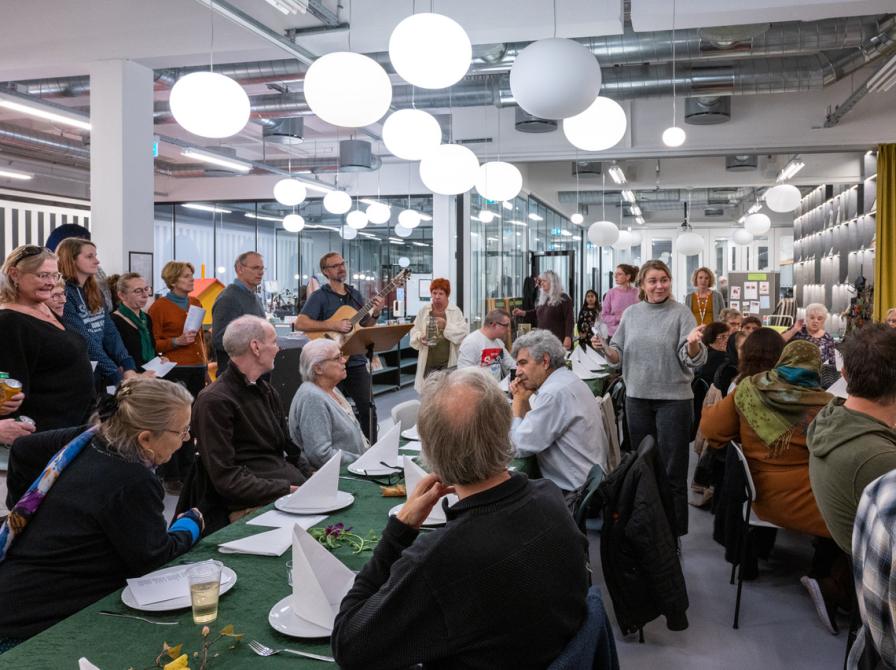 Foto af mange forskellige mennesker, der mødes til fællesspisning og møde. En kvinde holder velkomsttale i midten af forsamlingen og alles opmærksomhed er rettet mod hende.