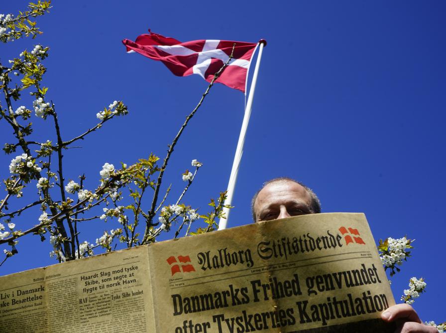 Foto af Hans, der står med en gammel avis fra Aalborg Stiftstidende, hvor man kan se, at forsiden handler om Danmarks Befrielse. Bag Hans er himlen smuk og blå, Dannebrog vejrer og et æbletræ blomstrer.