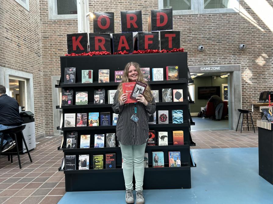 Sørine står foran Ordkraft udstillingen på Hovedbiblioteket. Udstillingen viser et stort udvalg af bøger skrevet af forfattere, der gæster festivalen i år. Sørine holder de to omtalte romaner i denne artikel i hænderne og smiler. Bag hende ses biblioteksrummet med bl.a. Gamezone.
